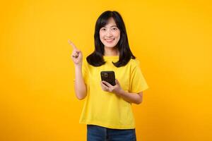 Capture young Asian woman wearing a yellow t-shirt and denim jeans points with finger hand gesture to free copy space while using smartphone. Perfect for illustrating the concept of app functionality. photo