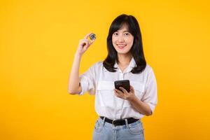 maravilloso retrato de un asiático joven mujer participación digital moneda y teléfono inteligente en amarillo estudio antecedentes. explorar el concepto de cripto digital divisa. foto