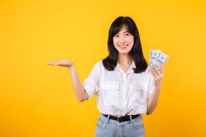 Young happy woman of Asian ethnicity wear white shirt and denim jean holding cash money in dollar and pointing hand to free copy space against yellow background. Investment and financial concept. photo