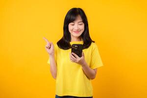 Capture young Asian woman wearing a yellow t-shirt and denim jeans points with finger hand gesture to free copy space while using smartphone. Perfect for illustrating the concept of app functionality. photo