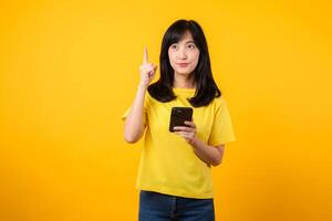 Capture young Asian woman wearing a yellow t-shirt and denim jeans points with finger hand gesture to free copy space while using smartphone. Perfect for illustrating the concept of app functionality. photo