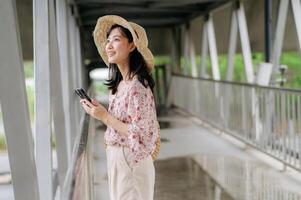 joven asiático mujer viajero con Costura cesta utilizando móvil teléfono y en pie en paso superior. viaje viaje estilo de vida, mundo viaje explorador o Asia verano turismo concepto. foto