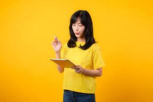 young Asian woman wearing yellow t-shirt and jeans showing happy smile while using digital tablet, displaying thoughtful expression and creative idea. education technology innovative thinking concept. photo