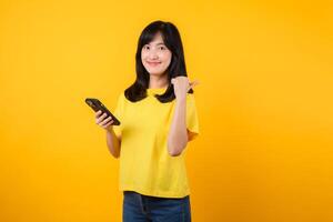 Capture young Asian woman wearing a yellow t-shirt and denim jeans points with finger hand gesture to free copy space while using smartphone. Perfect for illustrating the concept of app functionality. photo