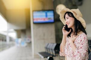 joven asiático mujer viajero con Costura cesta utilizando un móvil teléfono y esperando para tren en tren estación. viaje viaje estilo de vida, mundo viaje explorador o Asia verano turismo concepto. foto