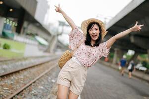 joven asiático mujer viajero con Costura cesta contento sonriente mirando a un cámara junto a tren ferrocarril. viaje viaje estilo de vida, mundo viaje explorador o Asia verano turismo concepto. foto