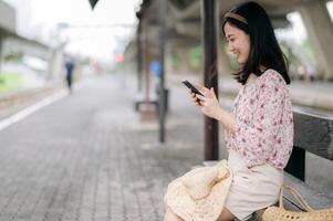 young asian woman traveler with weaving basket using a mobile phone and waiting for train in train station. Journey trip lifestyle, world travel explorer or Asia summer tourism concept. photo