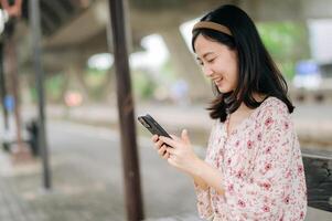 young asian woman traveler with weaving basket using a mobile phone and waiting for train in train station. Journey trip lifestyle, world travel explorer or Asia summer tourism concept. photo