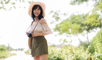 retrato de asiático joven mujer viajero con Costura sombrero y cesta y un cámara en verde público parque naturaleza antecedentes. viaje viaje estilo de vida, mundo viaje explorador o Asia verano turismo concepto. foto