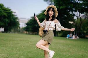 retrato de asiático joven mujer viajero con Costura sombrero, cesta, móvil teléfono y cámara en verde público parque antecedentes. viaje viaje estilo de vida, mundo viaje explorador o Asia verano turismo concepto. foto