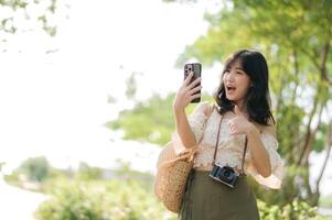 Portrait of asian young woman traveler with weaving hat, basket, mobile phone and camera on green public park background. Journey trip lifestyle, world travel explorer or Asia summer tourism concept. photo