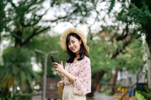 Portrait of asian young woman traveler with weaving hat and basket using mobile phone on green park nature background. Journey trip lifestyle, world travel explorer or Asia summer tourism concept. photo