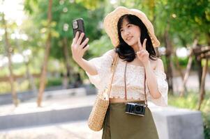 retrato de asiático joven mujer viajero con Costura sombrero, cesta, móvil teléfono y cámara en verde público parque antecedentes. viaje viaje estilo de vida, mundo viaje explorador o Asia verano turismo concepto. foto