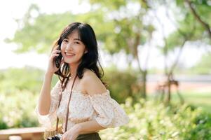 retrato de asiático joven mujer viajero con Costura sombrero, cesta, móvil teléfono y cámara en verde público parque antecedentes. viaje viaje estilo de vida, mundo viaje explorador o Asia verano turismo concepto. foto