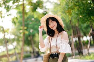 Portrait of asian young woman traveler with weaving hat and basket and a camera on green public park nature background. Journey trip lifestyle, world travel explorer or Asia summer tourism concept. photo