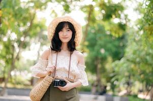 Portrait of asian young woman traveler with weaving hat and basket and a camera on green public park nature background. Journey trip lifestyle, world travel explorer or Asia summer tourism concept. photo