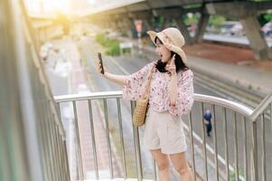 young asian woman traveler with weaving basket using mobile phone and standing on overpass with railway background. Journey trip lifestyle, world travel explorer or Asia summer tourism concept. photo