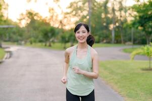 ajuste asiático joven mujer trotar en parque sonriente contento corriendo y disfrutando un sano al aire libre estilo de vida. hembra persona que practica jogging. aptitud corredor niña en público parque. sano estilo de vida y bienestar siendo concepto foto