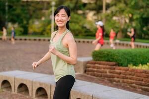 Female jogger. Fit young Asian woman with green sportswear aerobics dance exercise in park and enjoying a healthy outdoor. Fitness runner girl in public park. Wellness being concept photo