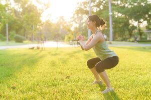 Female jogger. Fit young Asian woman with green sportswear squatting in park before running and enjoying a healthy outdoor. Fitness runner girl in public park. Wellness being concept photo