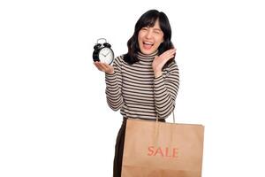 Portrait of excited young Asian woman with sweater shirt holding alarm clock and shopping paper bag isolated on white background. last deals shocked price shopping concept photo