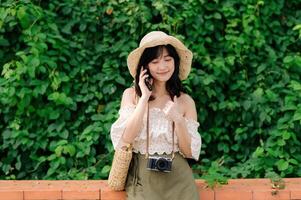 retrato de asiático joven mujer viajero con Costura sombrero, cesta, móvil teléfono y cámara en verde público parque antecedentes. viaje viaje estilo de vida, mundo viaje explorador o Asia verano turismo concepto. foto