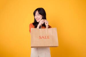 Portrait happy attractive 30s asian young woman with trendy springtime dress, orange shirt fashion and paper bag isolated on yellow background. Summertime sale shopping concept. photo