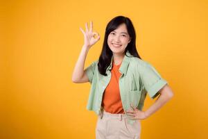 Embodying confidence and cheerful 30s Asian woman wearing green and orange shirt. With hand on hip and okay sign against yellow background, radiates positivity and self-assurance. Approved concept. photo