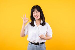 Young businesswoman hand sign okay gesture. Asian woman happy smile wearing white shirt and jean denim plants showing okay sign hand gesture isolated on yellow background. photo
