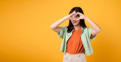 Asian young woman 30s wearing green and orange shirt. With a heart hand gesture on eye, adds whimsical touch. Unique and creative expression of affection on a vibrant yellow background. photo