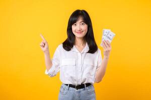 Young happy woman of Asian ethnicity wear white shirt and denim jean holding cash money in dollar and pointing finger to free copy space against yellow background. Investment and financial concept. photo