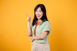 portrait joyful young asian woman model showing pointing finger gesture to free copy space isolated on yellow studio background. female adult posing for advertising. offer marketing concept. photo