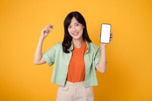 Happy asian young 30s woman wearing orange shirt green shirt holding digital crypto currency coin and smartphone isolated on yellow background. Digital currency financial concept. photo