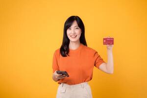 retrato alegre joven 30s asiático mujer contento sonrisa participación crédito tarjeta para compra en línea compras pago en teléfono inteligente aislado en amarillo antecedentes. foto