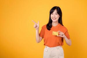 cheerful asian young woman 30s wearing orange shirt holding credit card and pointing finger towards free copy space isolated on yellow background. positivity, shopping, and financial concepts. photo