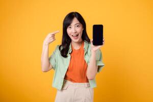 alegre asiático mujer 30s vistiendo naranja y verde camisa. puntos dedo a teléfono inteligente, simbolizando conectividad y digital empoderamiento. tecnología y moderno estilo de vida en un vibrante amarillo antecedentes. foto