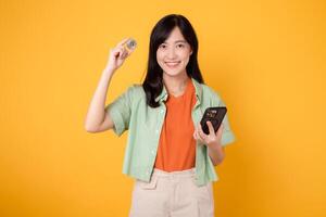 Asian woman 30s holding crypto currency coin while using smartphone, embodies the world of financial innovation and digital currency. technology and finance on yellow background. photo