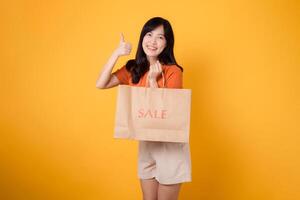 Portrait happy attractive 30s asian young woman with trendy springtime dress, orange shirt fashion and paper bag isolated on yellow background. Summertime sale shopping concept. photo
