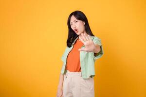 cheerful Asian woman 30s wearing green and orange shirt. With a hand raised in 'no' gesture, expresses conviction isolated on yellow background. photo