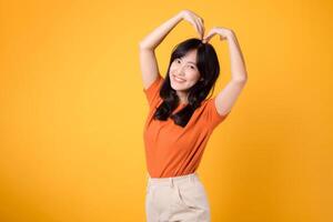 Spread love and joy with cheerful Asian woman in 30s wearing orange shirt show happy smile while forming heart shape with arms, symbolizing love and romance. Illustrating love and romantic concepts. photo