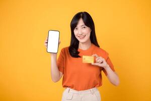 retrato alegre 30s joven asiático mujer contento sonrisa participación crédito tarjeta para compra en línea compras pago y demostración blanco teléfono inteligente pantalla monitor para publicidad aislado en amarillo antecedentes. foto