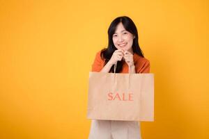 Cheerful Asian woman 30s wearing orange shirt showing happy smile while proudly displaying sale paper bag against yellow background. shopping and enjoying great deals concept. photo