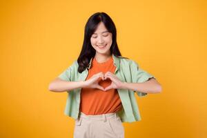 portrait grateful joy young asian woman making heart hands shape to her chest isolated on yellow background. expression peaceful, thankful emotion happiness love positive feeling. photo