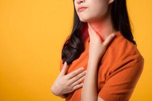 Expressing care wellness, cheerful young Asian woman in 30s wearing orange shirt holding her throat, portraying sore throat. Perfect for representing health concerns and the importance of self-care. photo