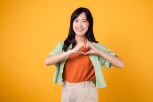 Cheerful Asian woman 30s wearing orange shirt showing heart hand sign gesture on chest against yellow background, symbolizing love and positivity. happiness and spreading heartfelt emotions concept. photo