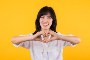 Young female healthcare. Asian woman wearing white shirt feels happy and romantic shapes heart gesture expresses tender feeling poses isolated on yellow background. People affection and care concept. photo