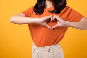 Close up of Asian woman 30s wearing orange shirt showing heart hand gesture, symbolizing affection and warmth. Perfect for expressing love and spreading positive vibes in various projects. photo