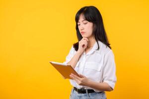 Doubtful Asian young adult business woman working on digital tablet and looking away thinking of solution isolated on yellow background, considering offer, making decision or feeling doubt concept. photo