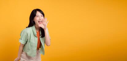 Vibrant young Asian woman in her 30s wearing a green and orange shirt, energetically shouting with her mouth isolated on a yellow background. Explore the discount shopping promotion concept. photo