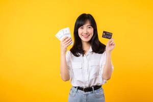 happy successful confident young asian woman happy smile wearing white shirt and denim jean holding cash money and credit card standing over yellow background. millionaire business, shopping concept. photo